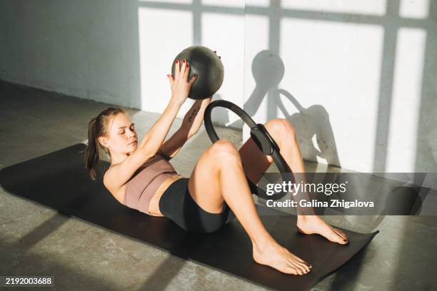 young slim woman in sportswear doing pilates stretching on mat with ring and ball in modern studio - cerchio in metallo da pilates foto e immagini stock