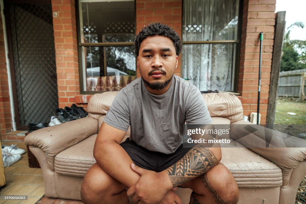 Tattooed Pacific Islander man posing for camera while sitting on a sofa in front of his house