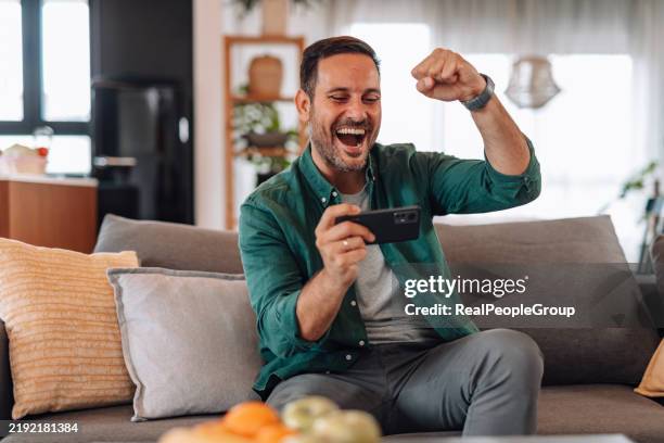 hombre emocionado ganando en un juego móvil en el sofá de casa - jugar a juegos de azar fotografías e imágenes de stock