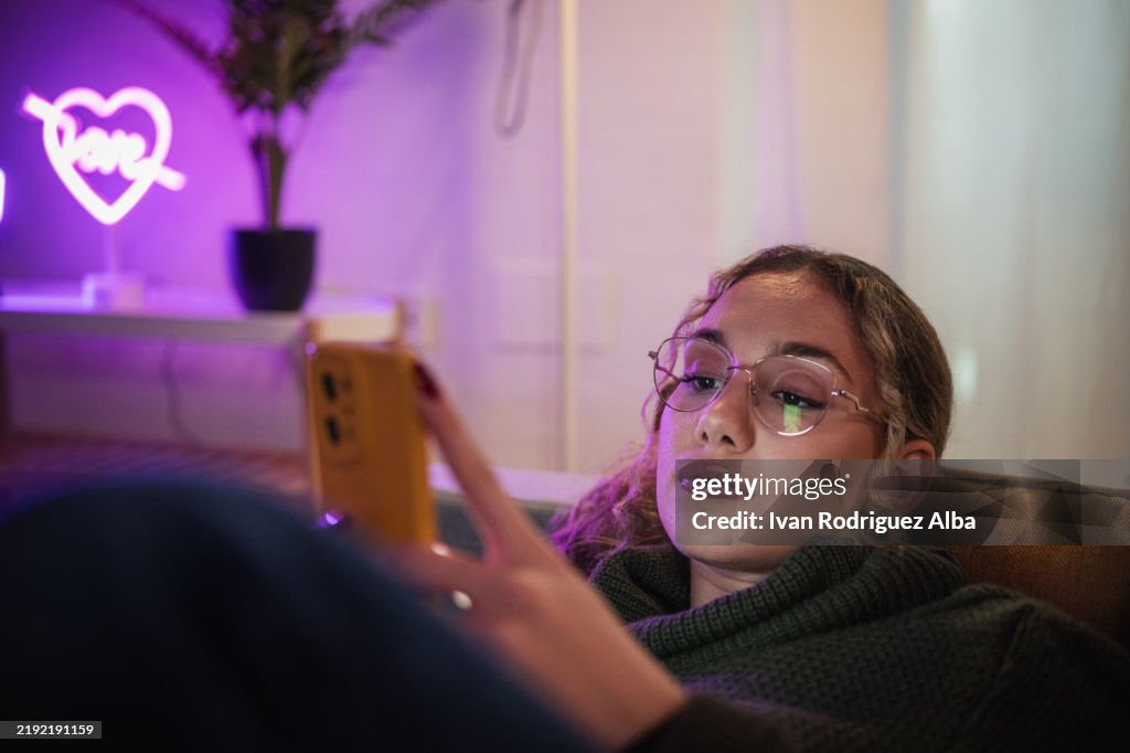 Young woman relaxing on sofa using smartphone in neon-lit room