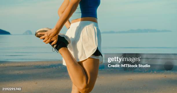 closeup of young asian woman in sportswear stretching on beach, preparing for jog along serene coastline in morning sunlight. exercise and healthy lifestyle. - female quads stock pictures, royalty-free photos & images