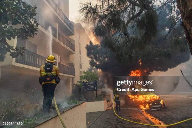 Firefighters sprays water towards a car and a complex building as flames from the Palisades Fire burn on Sunset Boulevard during a powerful windstorm...