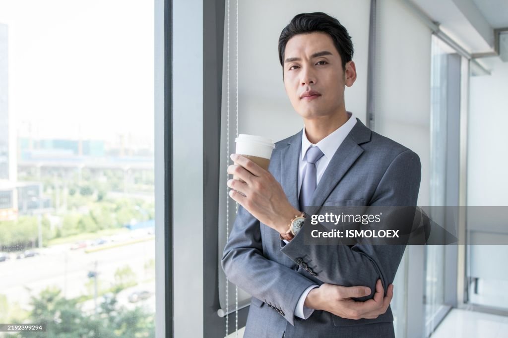 Businessman in suit standing by window holding coffee cup looking at camera