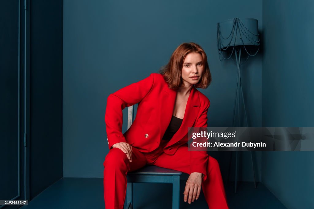 Portrait looking at camera serious fashion woman in red suit sitting in chair looking at camera