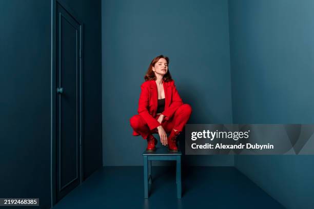 fashionable woman in red suit posing squatting on stool studio shot. conception fashion - postura foto e immagini stock