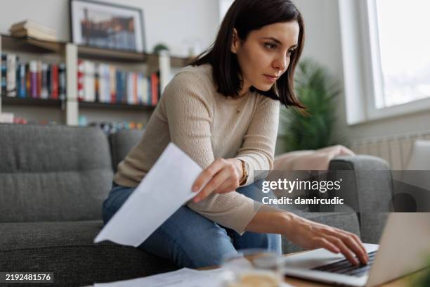 donna che esamina i documenti finanziari mentre lavora sul suo laptop a casa - paragone foto e immagini stock