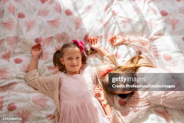 two young girls lying on a floral-patterned bedspread enjoying playful company - pale pink stock pictures, royalty-free photos & images