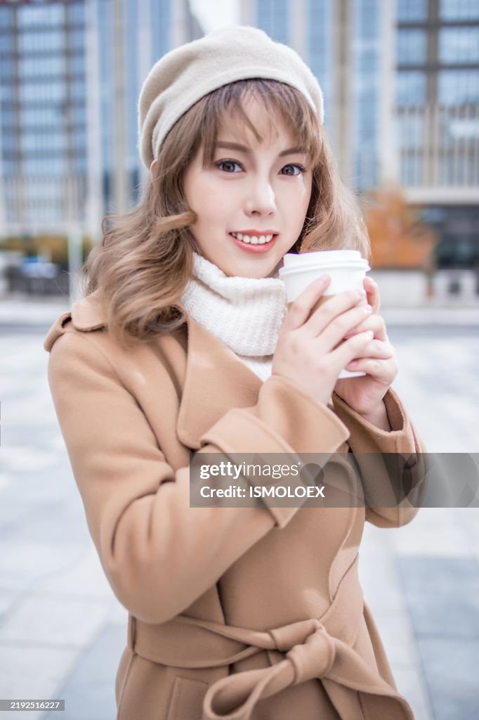 Eine junge Frau in einem braunen Mantel und cremefarbener Baskenmütze lächelt, während sie eine Tasse Kaffee vor einem modernen Gebäude hält