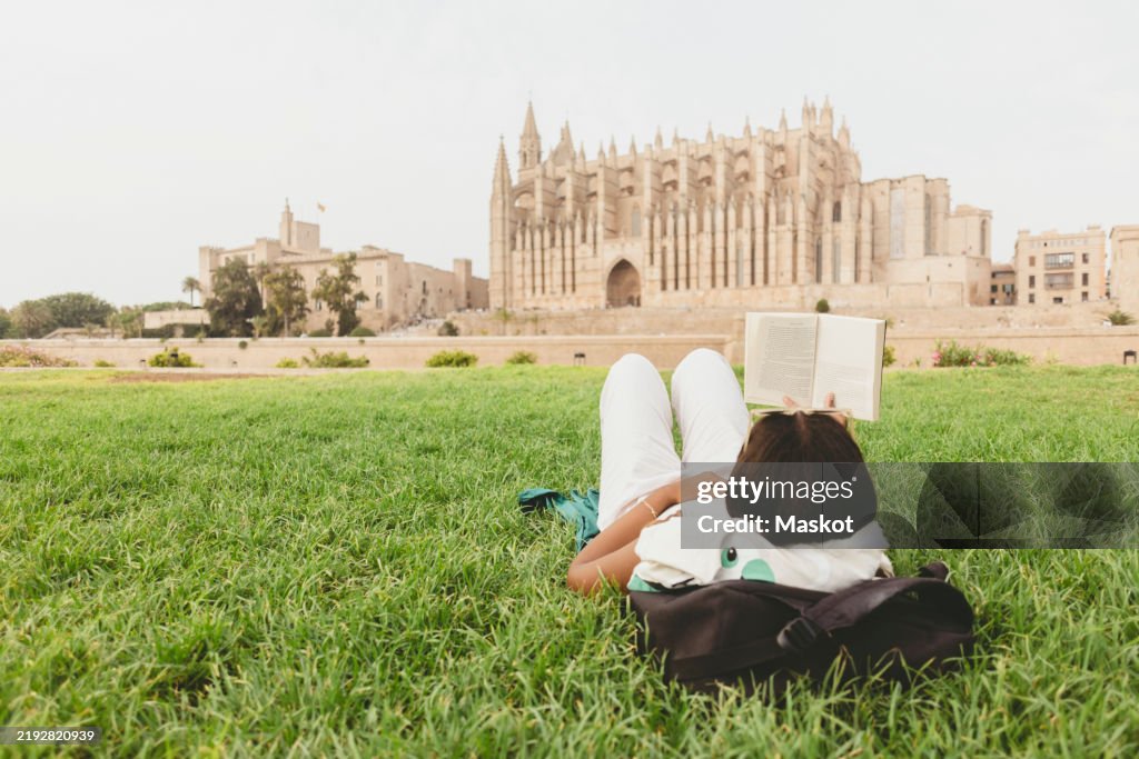 Female tourist reading book while lying down on grass against Palma Cathedral