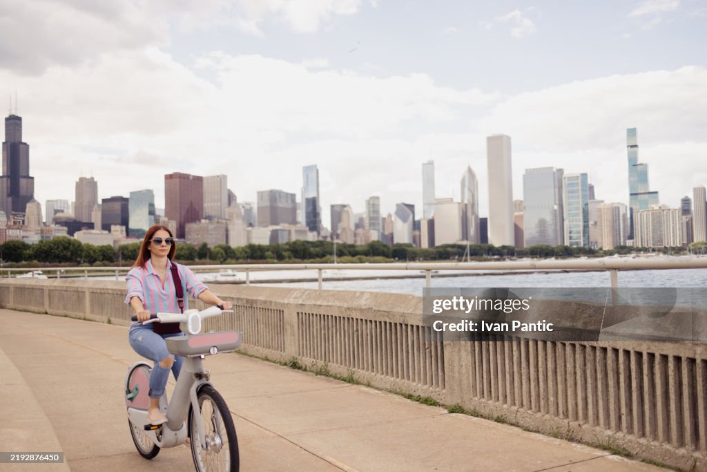Woman Riding Bike Along Lakeside Path With Cityscape in Background