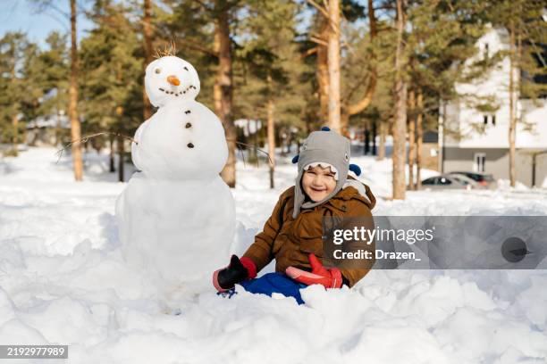 child posing while building a snowman in a snow-covered neighborhood during a sunny winter day on christmas day - snowman building stock pictures, royalty-free photos & images