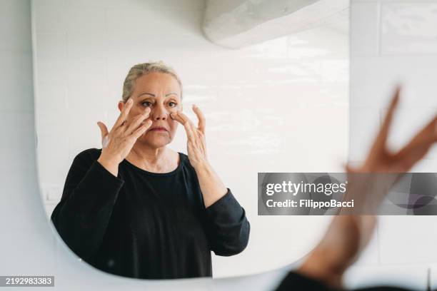 seniorin trägt anti-aging-creme auf das gesicht auf, das in den spiegel schaut - anti age stock-fotos und bilder