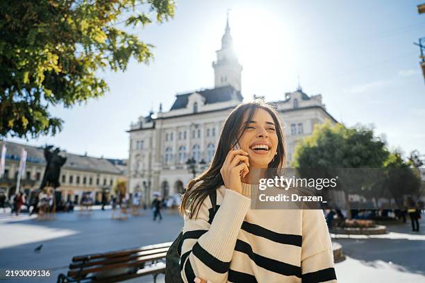 young woman laughing while talking on smartphone in sunny city square with historic building in the background - market square stock pictures, royalty-free photos & images