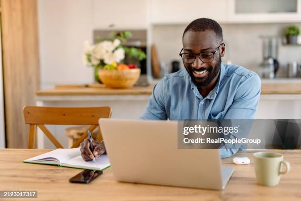 uomo felice che lavora da casa prendendo appunti guardando il computer portatile - educazione online foto e immagini stock