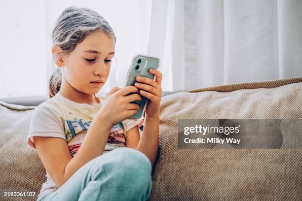 cute little girl using technology device sitting on sofa. girl playing on phone. - miopia foto e immagini stock