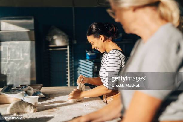 smiling female baker kneading dough with senior colleague while working at bakery - baking bread stock pictures, royalty-free photos & images