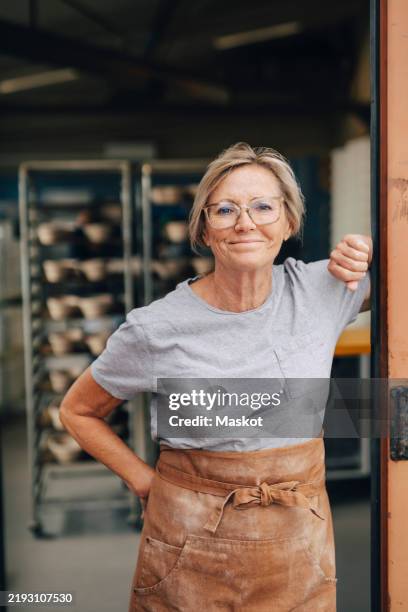 confident senior female baker standing with hand on hip at bakery doorway - retraités qui travaillent photos et images de collection