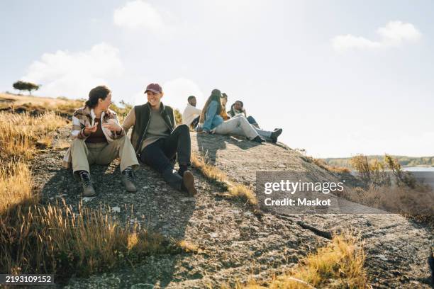 happy young man talking with female friend while sitting on hill at sunny day - svensk sommar bildbanksfoton och bilder