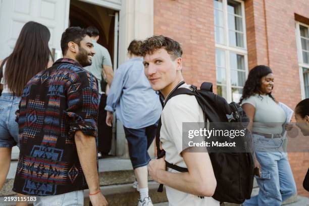 portrait of smiling man carrying bag and moving up stairs while looking over shoulder at college campus - nach oben gucken stock-fotos und bilder