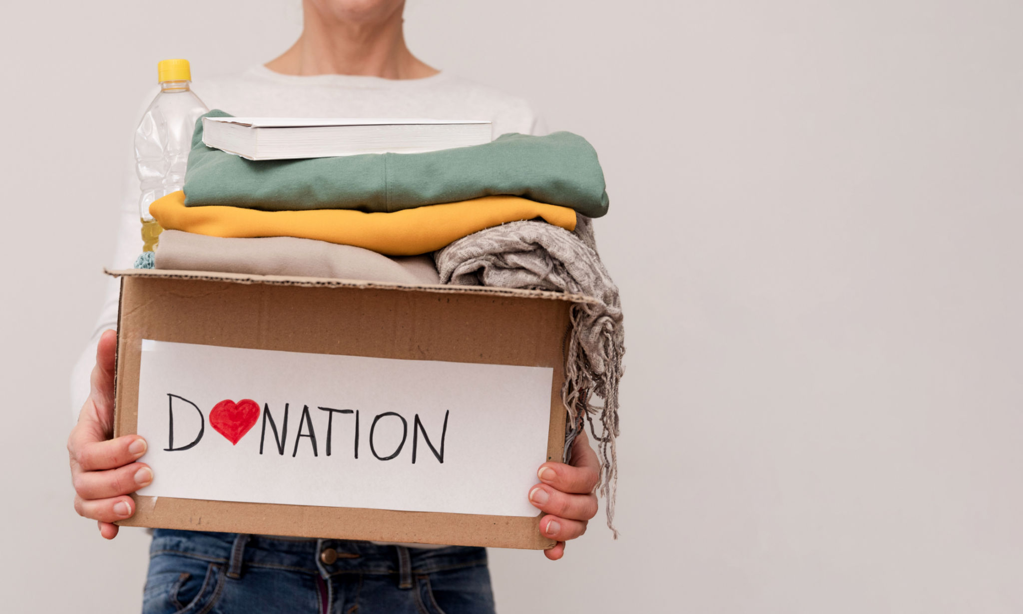 Close up of Volunteer holding donation box with clothes, food and book on grey background with copy space Close up of Volunteer holding donation box with clothes, food and book on grey background with copy space