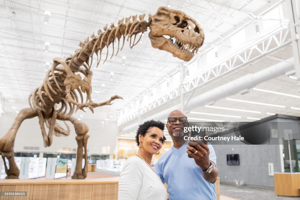 A retired couple takes a selfie with a T-rex fossil in the background.
