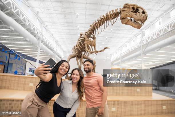 in a library lobby, a group of three friends pose for a photo with a dinosaur fossil behind them. - t rex museum stock pictures, royalty-free photos & images