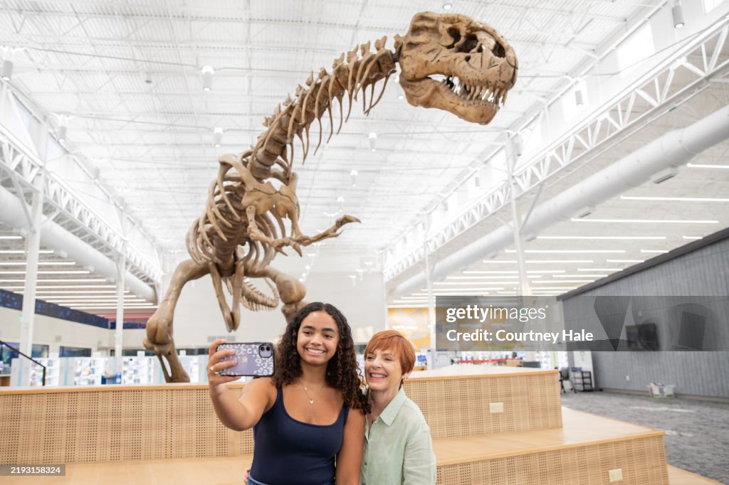 A teen girl and her mom pose for a selfie in front of a dinosaur fossil display.