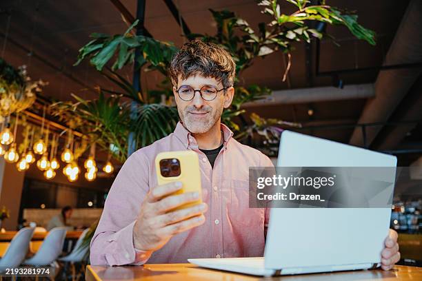 businessman seated at a cozy cafe table engaged in a phone call and laptop with ambient warm lighting providing a comfortable setting - financial occupation stock pictures, royalty-free photos & images