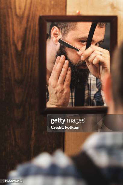 a young and handsome man with a beard begins to shave in front of a mirror. - wood shaving stock pictures, royalty-free photos & images