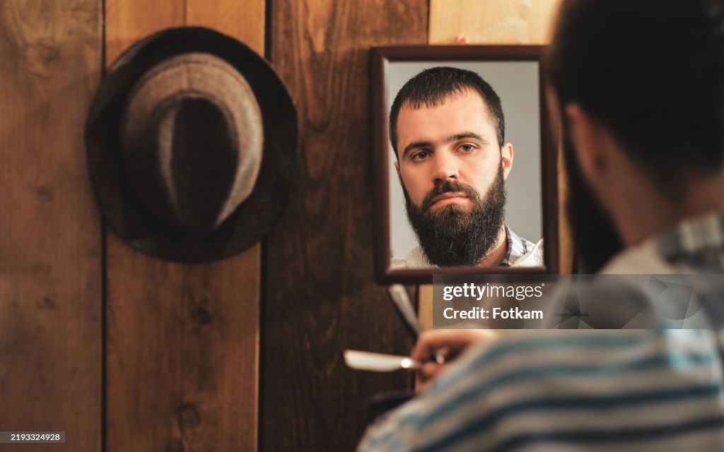 Young and handsome man with a beard holds a Straight Razor in his hand for shaving in front of a mirror.