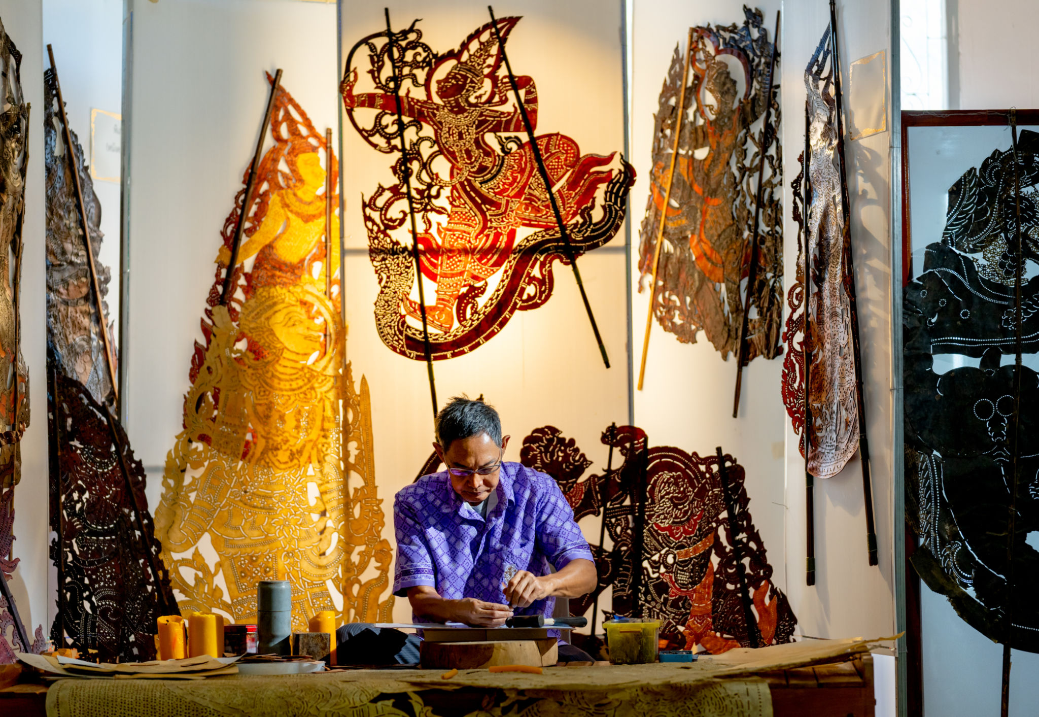 Front view Asian senior man wear glasses use hammer and chisel to create grand shadow play craft and he concentrate to work in area of workplace with several products are place around workplace area. Front view Asian senior man wear glasses use hammer and chisel to create grand shadow play craft and he concentrate to work in area of workplace with several products are place around workplace area.