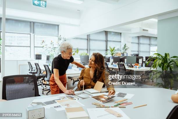 two designers collaborating at a brightly lit office desk reviewing documents and discussing creative ideas together - design studio stock pictures, royalty-free photos & images