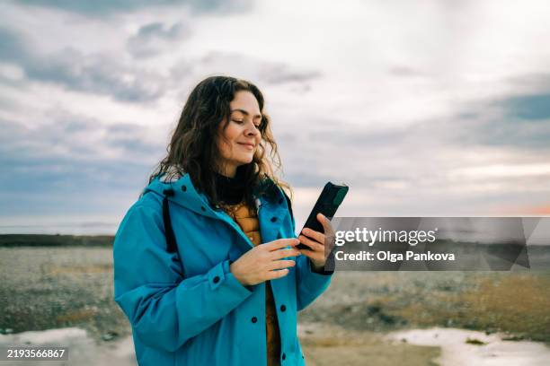 woman wearing a blue raincoat using a smartphone on a cold beach at sunset - rainwear stock pictures, royalty-free photos & images