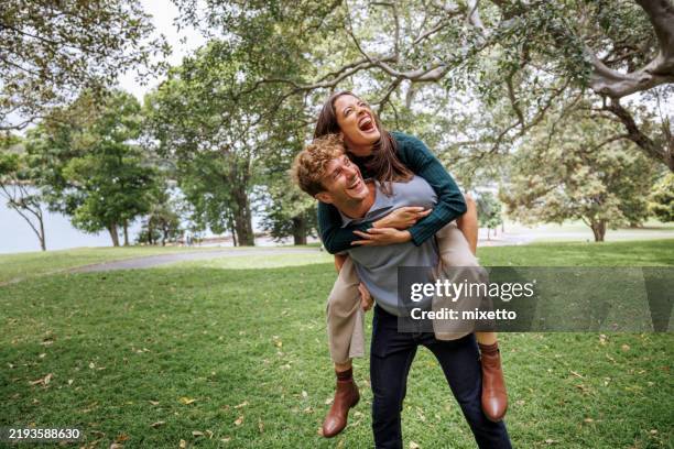 cheerful young man giving piggyback ride to his girlfriend outdoors at the public park - man giving girlfriend piggy back ride in park stock pictures, royalty-free photos & images