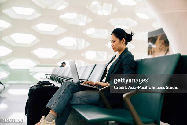 asian business woman working on laptop while sitting at airport terminal waiting for her flight, a suitcase by her side. business traveller. lifestyle and technology. business and leisure travel. business on the go. - business class imagens e fotografias de stock
