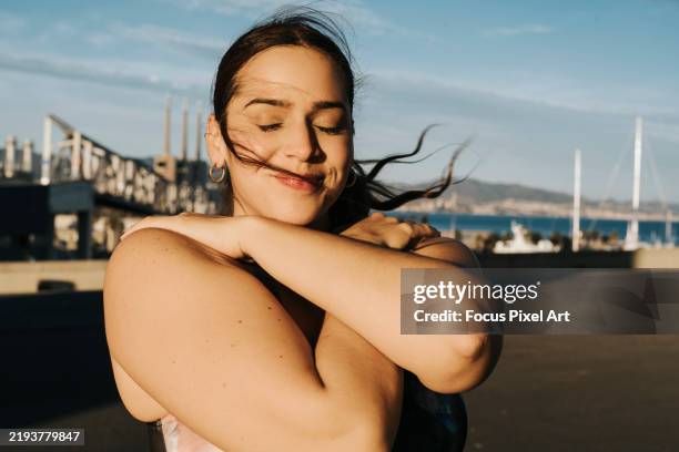 peaceful female gently holding herself, eyes closed, embodying self-care amid urban landscape, radiating inner calm - voluptuoso fotografías e imágenes de stock