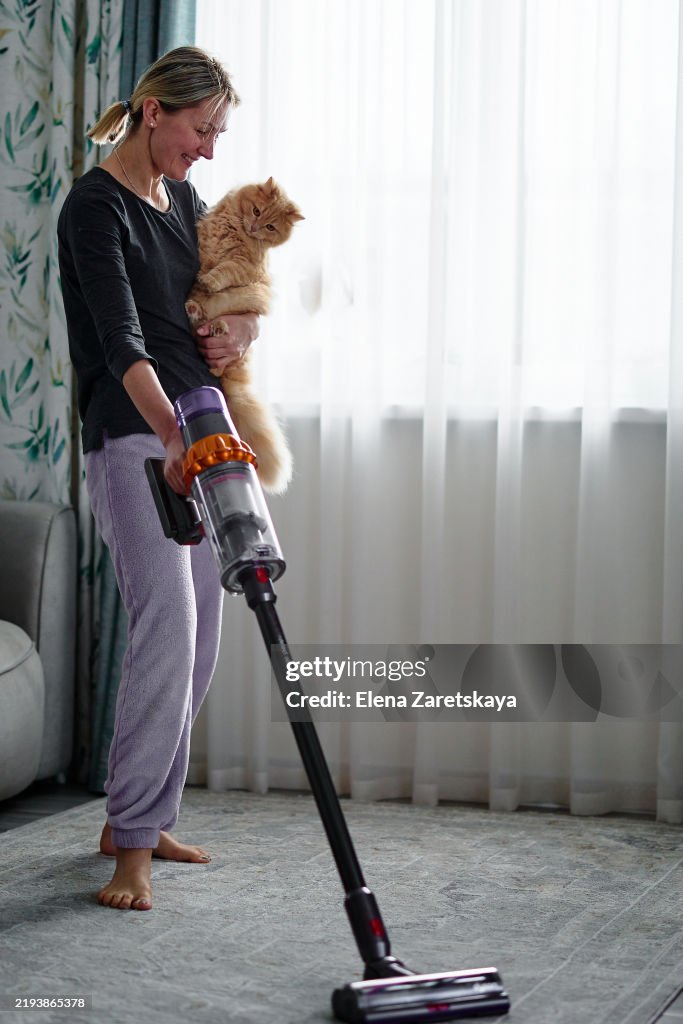 Woman Holding Cat While Vacuuming a Carpet in a Bright Living Room