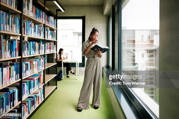 a stylish young female student stands in modern library with shelves, study desks, and large windows - librarian reading stock pictures, royalty-free photos & images