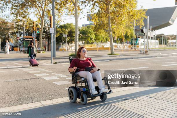 happy woman using electric wheelchair crossing street in the city - rolstoel stockfoto's en -beelden