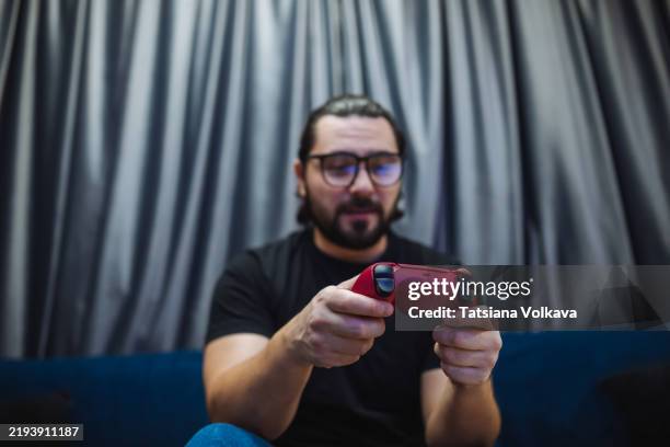 man in glasses focused while holding a red controller while on blue couch. gray curtains visible in the background. good for gaming content. - console controller stock pictures, royalty-free photos & images