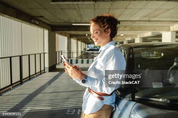smiling woman checking her phone near a car in a parking garage - parking stock pictures, royalty-free photos & images