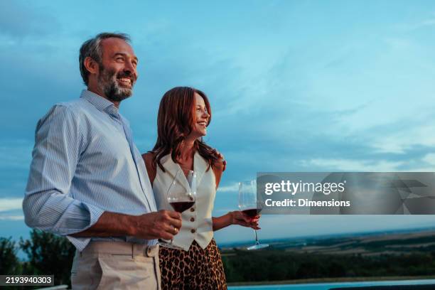 couple mature heureux buvant du vin au bord de la piscine à débordement au coucher du soleil - richesse photos et images de collection