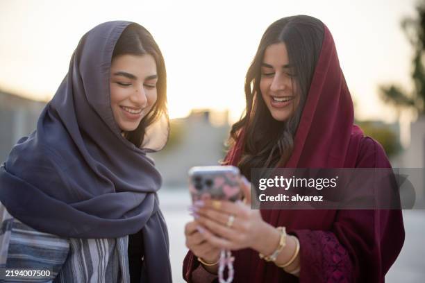 two middle eastern woman friends looking at mobile phone and smiling outside - middle eastern culture stock pictures, royalty-free photos & images