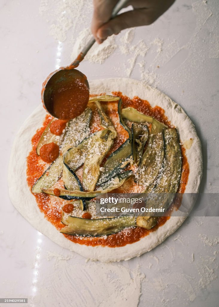 Close-up of a chef adding extra tomato sauce over grilled eggplant and grated cheese on raw pizza dough, traditional Italian pizza preparation in a professional pizzeria kitchen