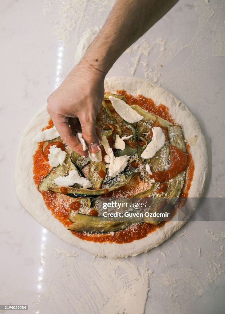 Close-up of a chef adding fresh mozzarella over grilled eggplant and tomato sauce on raw pizza dough, traditional Italian pizza preparation in a professional pizzeria kitchen