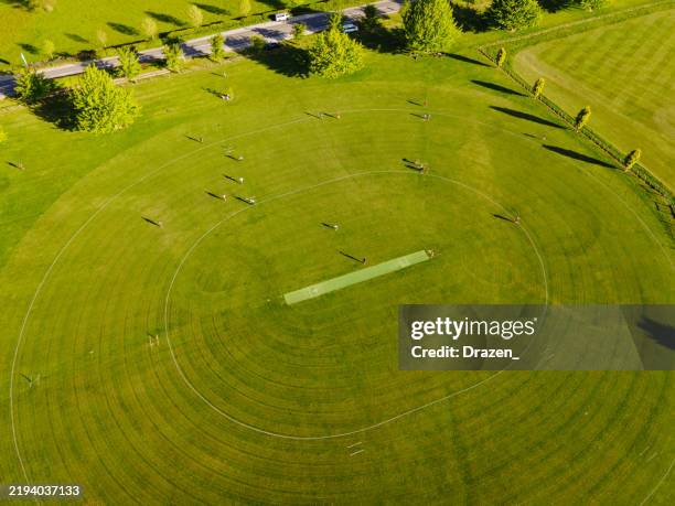 vue aérienne d’un terrain de cricket entouré d’arbres verdoyants avec des joueurs projetant de longues ombres par une journée ensoleillée - terrain-de-cricket photos et images de collection