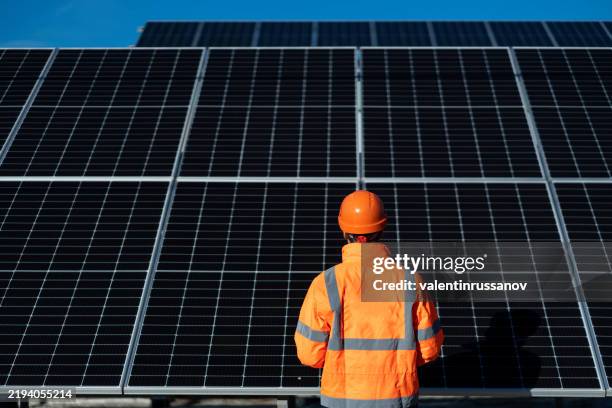 un ingeniero con chaqueta naranja inspecciona grandes paneles solares bajo un cielo azul claro como telón de fondo, un día soleado. - generadores fotografías e imágenes de stock