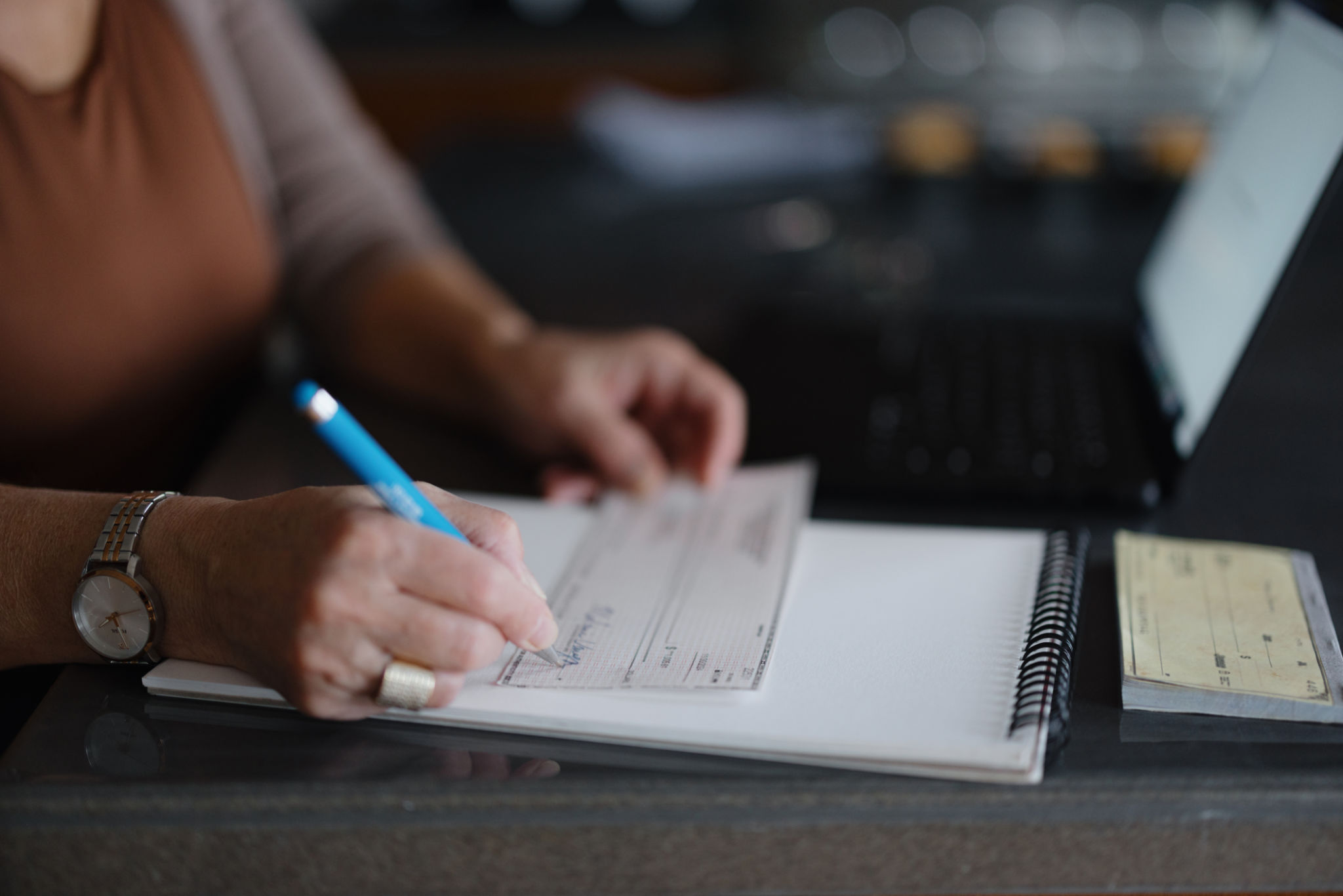 Woman Writing a Check at Desk with Notebook and Laptop Nearby Woman Writing a Check at Desk with Notebook and Laptop Nearby