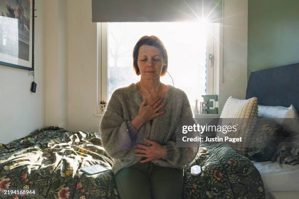 a mature woman uses a medical device for vagus nerve stimulation (neuromodulation) whilst sitting on her bed. a low winter sun streams uplifting light into the room - women stock pictures, royalty-free photos & images