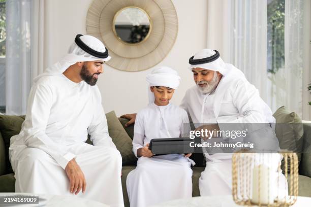 emirati boy holding digital tablet sitting between father and grandfather on couch, concentrating - gesturing stock pictures, royalty-free photos & images